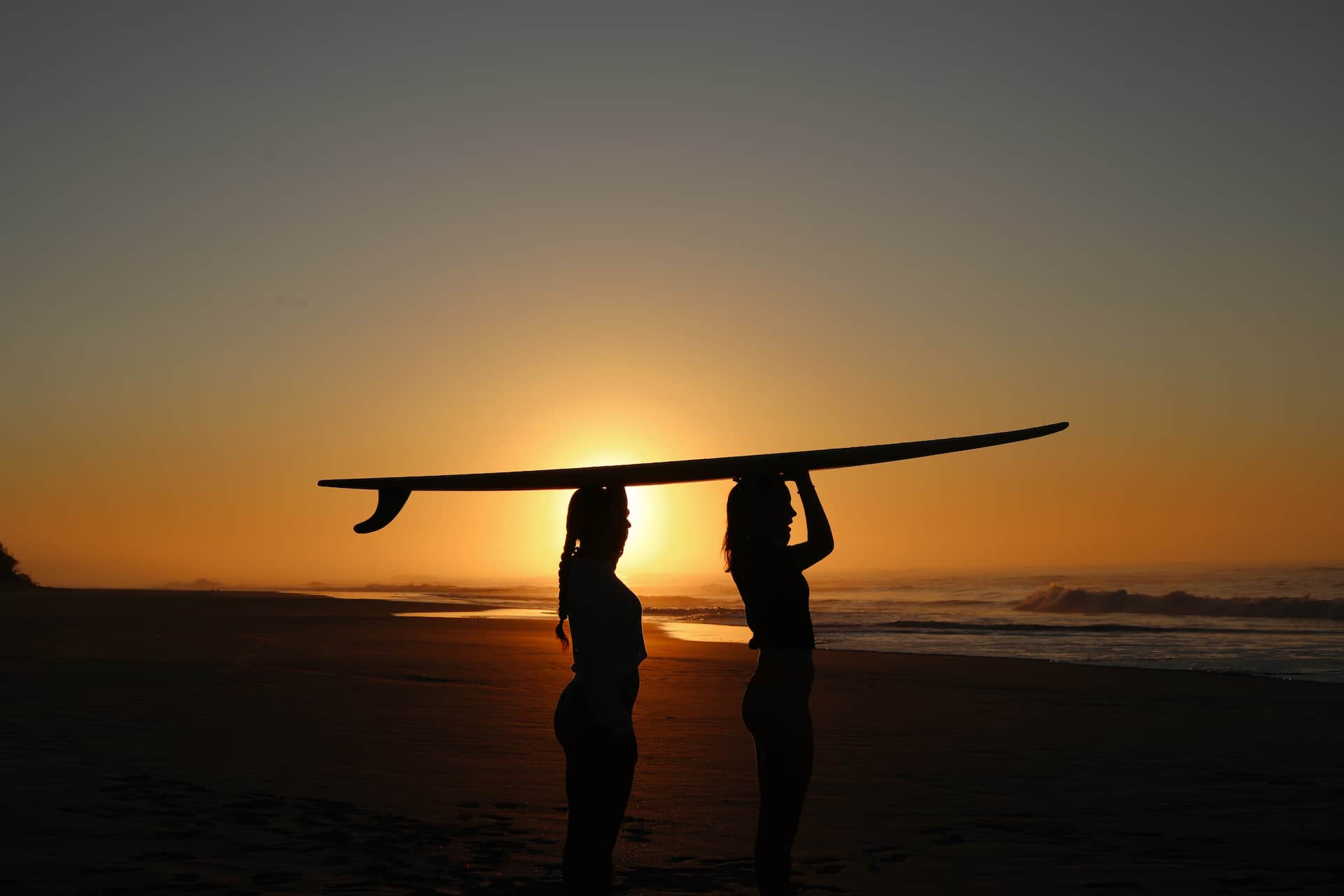 Two surfers carrying a surfboard along the beach at sunset